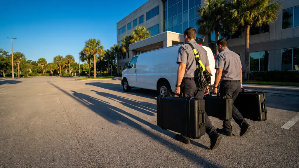 Two installers walk from a white van to an office building carrying equipment cases.