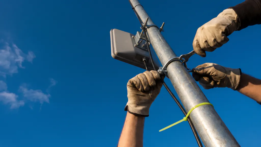 Gloved hands tighten a clamp on an antenna mast against a blue sky.