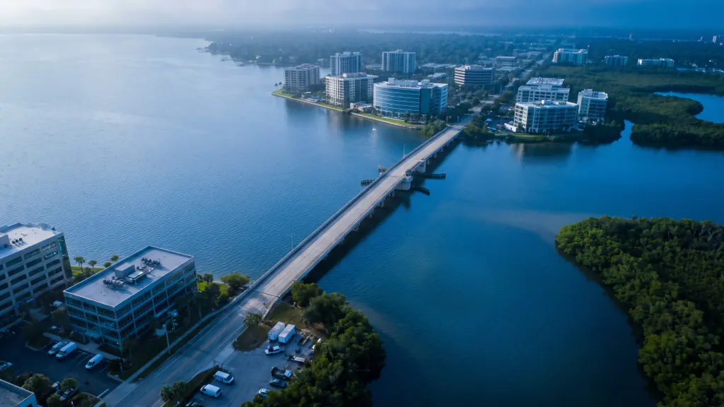 Aerial of a bay city with mid-rise buildings, a causeway and mangroves in morning haze.