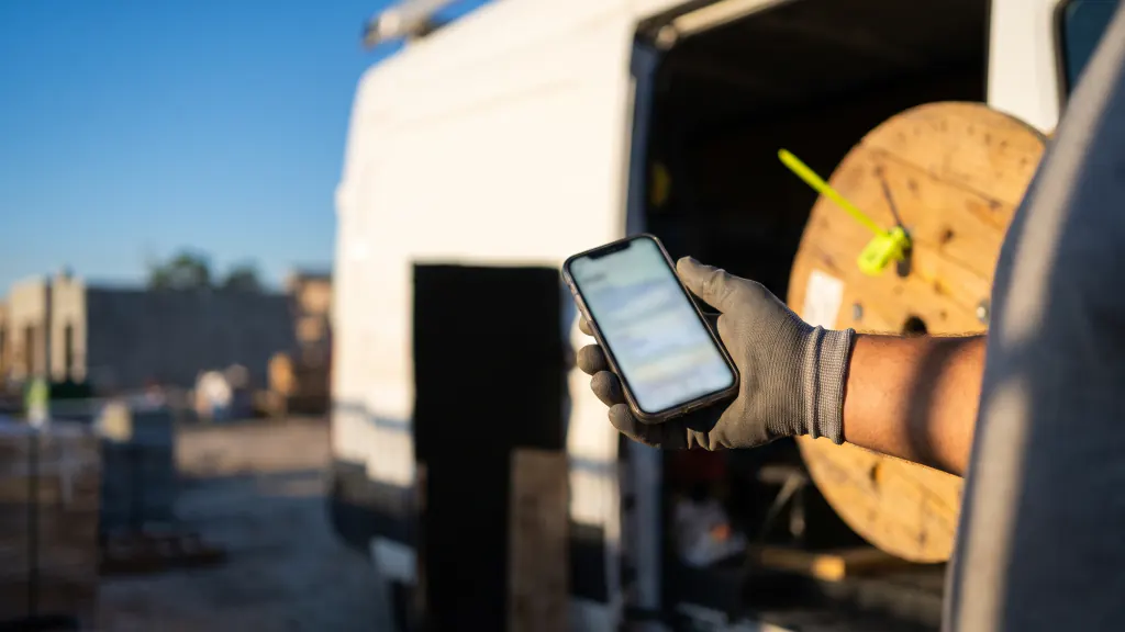 An installer's gloved hand holds a phone at a jobsite, a van and cable reel behind.