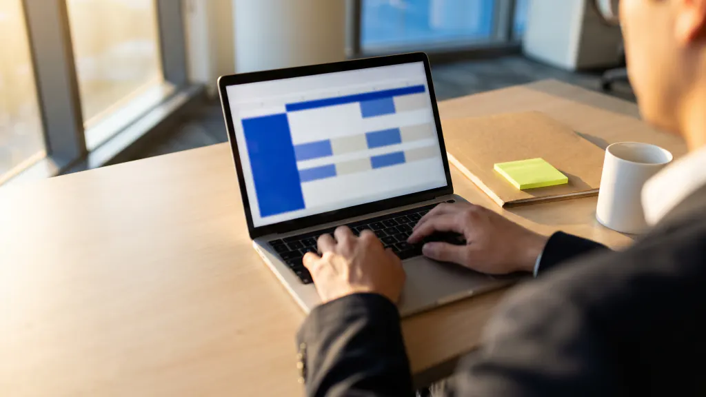 Hands on a laptop showing a blurred table of records, with a folder and coffee on the desk.